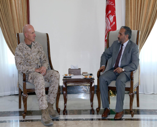 Two men in chairs facing each other across a table with plates, a tissue box, and other objects, in a formal setting with curtains and a flag in the background, engaged in conversation.