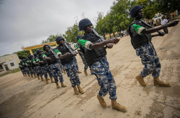 Nigerian soldiers in helmets and uniforms march down a dirt road carrying rifles, with trees, buildings, and a clear sky in the background; a vehicle and civilians stand to the right.
