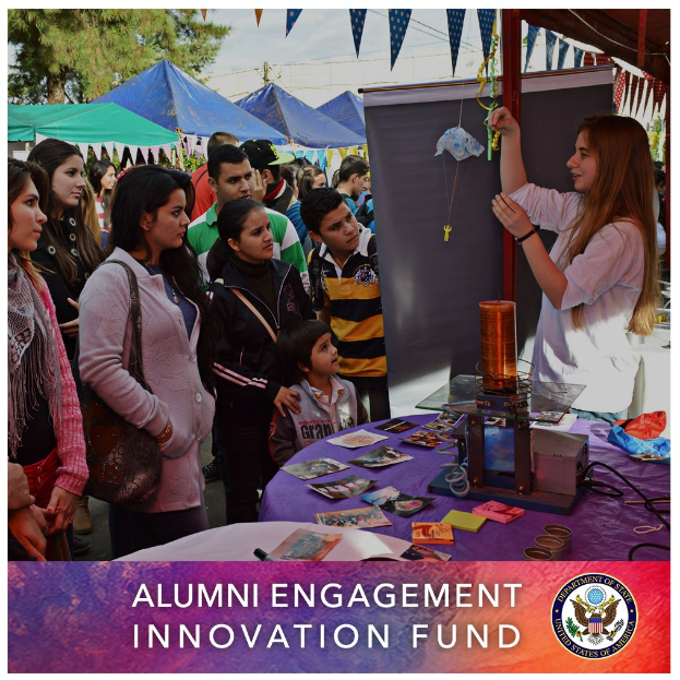 Gruppe von Menschen um einen Tisch versammelt mit einer "Alumni Engagement Innovation Fund"-Aufschrift, umgeben von Gegenständen, mit Zelten und Bäumen im Hintergrund unter einem klaren blauen Himmel, und Text und ein Logo unten.