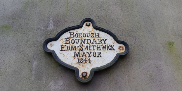 Schwarze Metalltafel an einem Gebäude mit weißer fettgedruckter Schrift, die "Gemeindegrenze Edm Smithwick Bürgermeister" lautet.