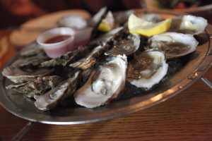 Ein Teller mit Garnelen in einer Reihe angeordnet, mit einer Tasse Sauce in der Mitte, zwei Zitronenstücken daneben und zusätzlichen Tellern im Hintergrund auf einem Tisch.