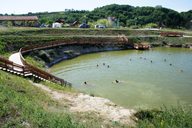 Menschen schwimmen in einem Gewässer umgeben von Grün, mit einer Brücke und Treppen zum Wasser, Schuppen, Fahrzeugen und Pfählen im Hintergrund unter einem klaren blauen Himmel.
