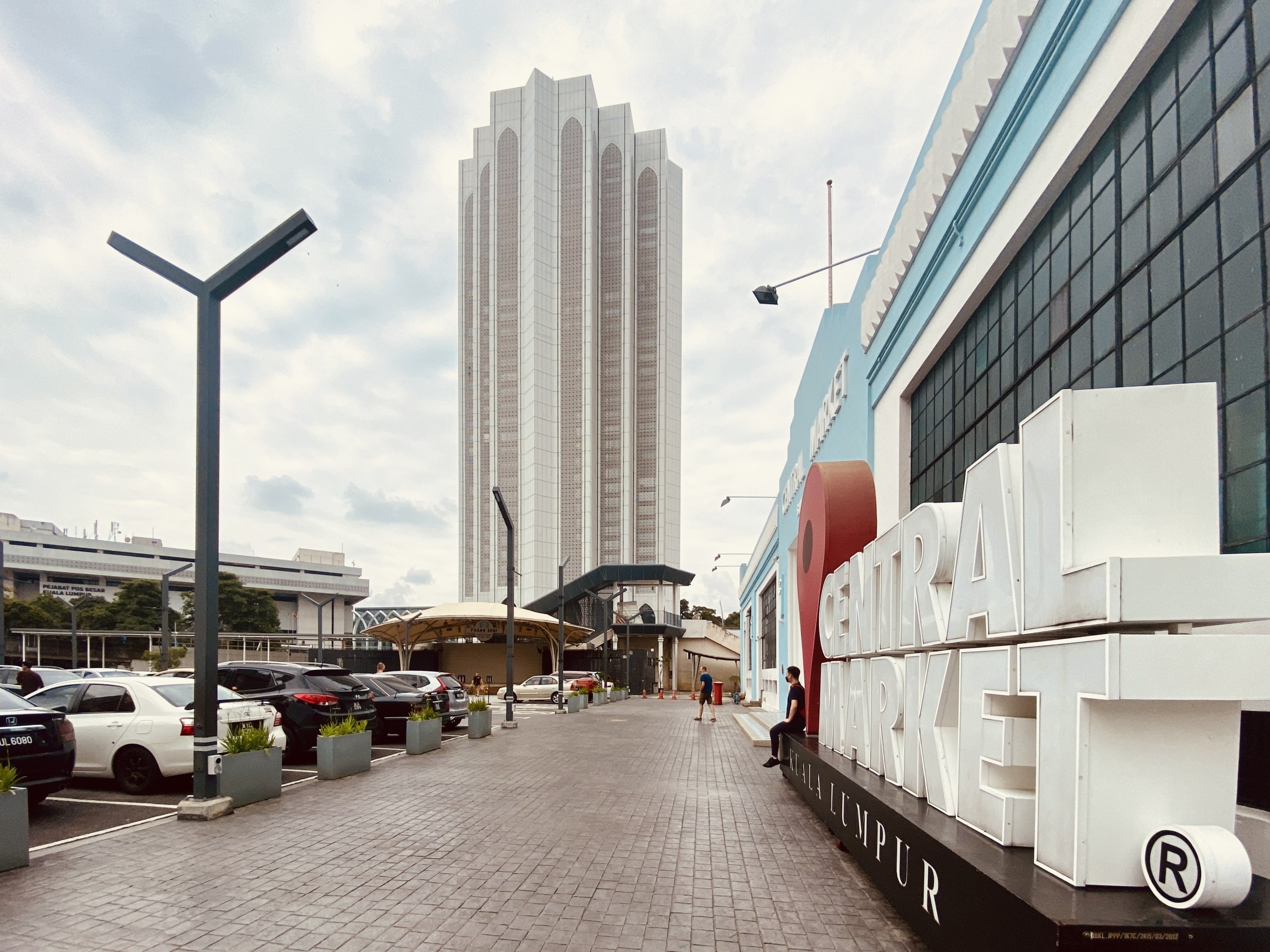 A busy central market in Kuala Lumpur, Malaysia, featuring tall buildings, street poles, vehicles, pedestrians, signboards, trees, and a cloudy sky.