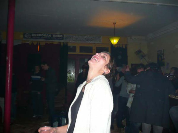 Eine Frau hält ein Glas und steht vor einer Gruppe von Menschen, Vorhängen, Fenstern, einer Wand, einer Rohrleitung und einer Lampe im Hintergrund.