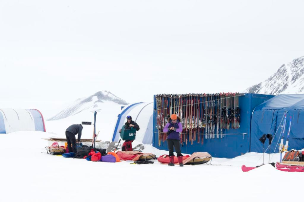 Drei Personen stehen auf einer verschneiten Landschaft mit Taschen, die herumliegen, Zelten mit Skiern dahinter und schneebedeckten Hügeln im Hintergrund unter einem klaren Himmel.
