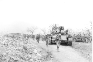 Ein Schwarz-Weiß-Bild von einer Gruppe von Menschen, die auf einer Straße neben einem Panzer gehen, mit anderen Menschen im Hintergrund, Steinen auf der linken Seite, Bäumen, einem Haus und dem Himmel darüber.