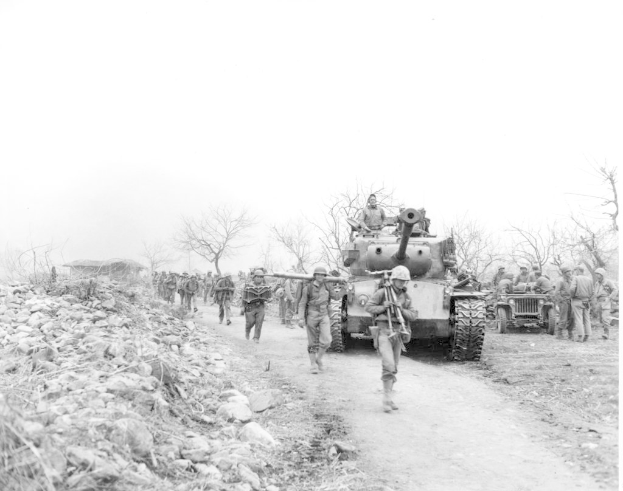 Ein Schwarz-Weiß-Bild von einer Gruppe von Menschen, die auf einer Straße neben einem Panzer gehen, mit anderen Menschen im Hintergrund, Steinen auf der linken Seite, Bäumen, einem Haus und dem Himmel darüber.