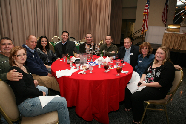 Eine Gruppe von Menschen mit einem freundlichen Gesichtsausdruck an einem Tisch mit Gläsern, Papieren, Weingläsern und Flaschen, einer Flagge auf der rechten Seite und Vorhängen an der Wand.