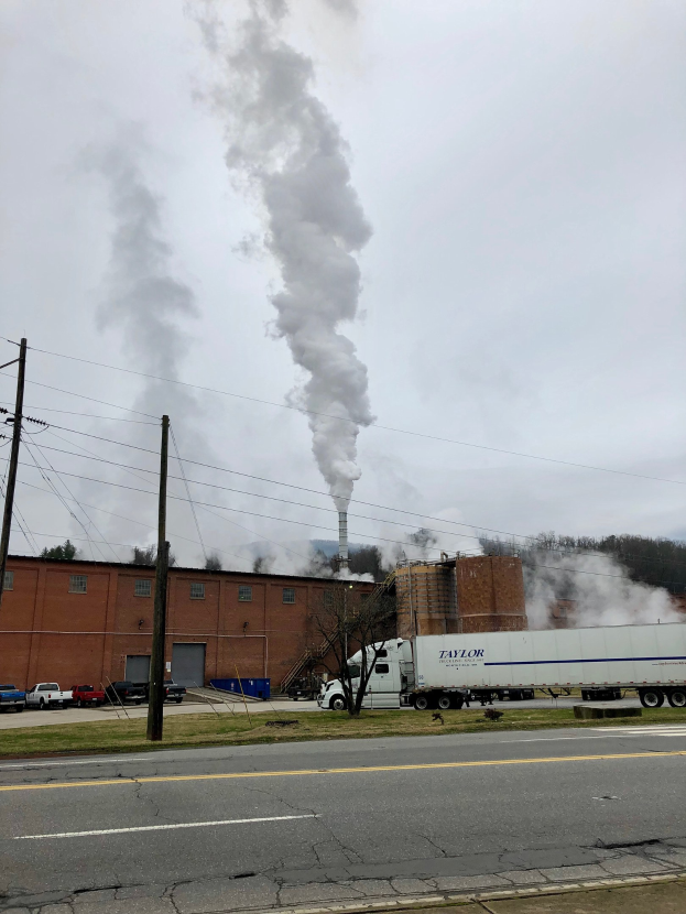 Ein Lastwagen fährt auf einer Straße mit einer im Hintergrund rauchenden Fabrik, umgeben von Gras, Bäumen und Strommasten.