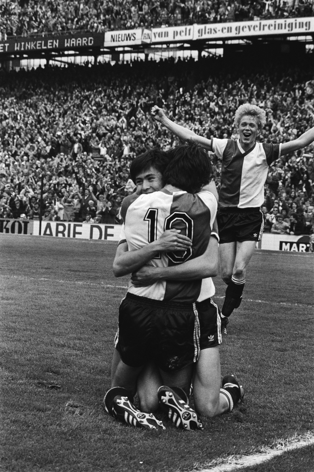 Drei Fussballspieler umarmen sich auf dem Boden und feiern ein Tor vor einer jubelnden Menge in einer schwarz-weissen Stadion-Szene.