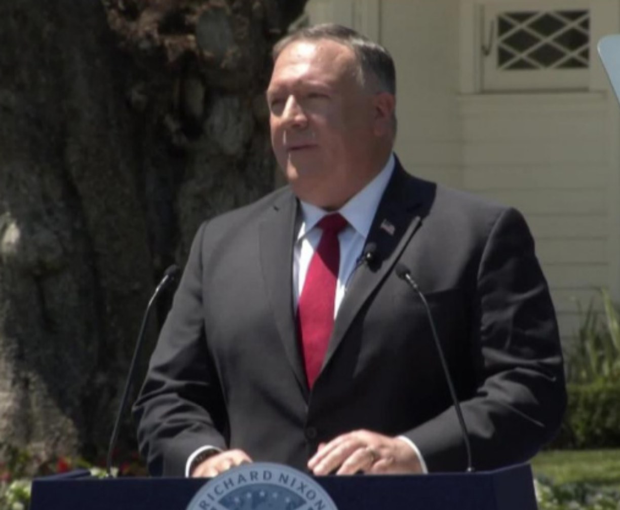Secretary of State Mike Pompeo stands at a podium with two microphones, delivering a speech with a tree, plants, and a building in the background; a board with text is visible on the podium.