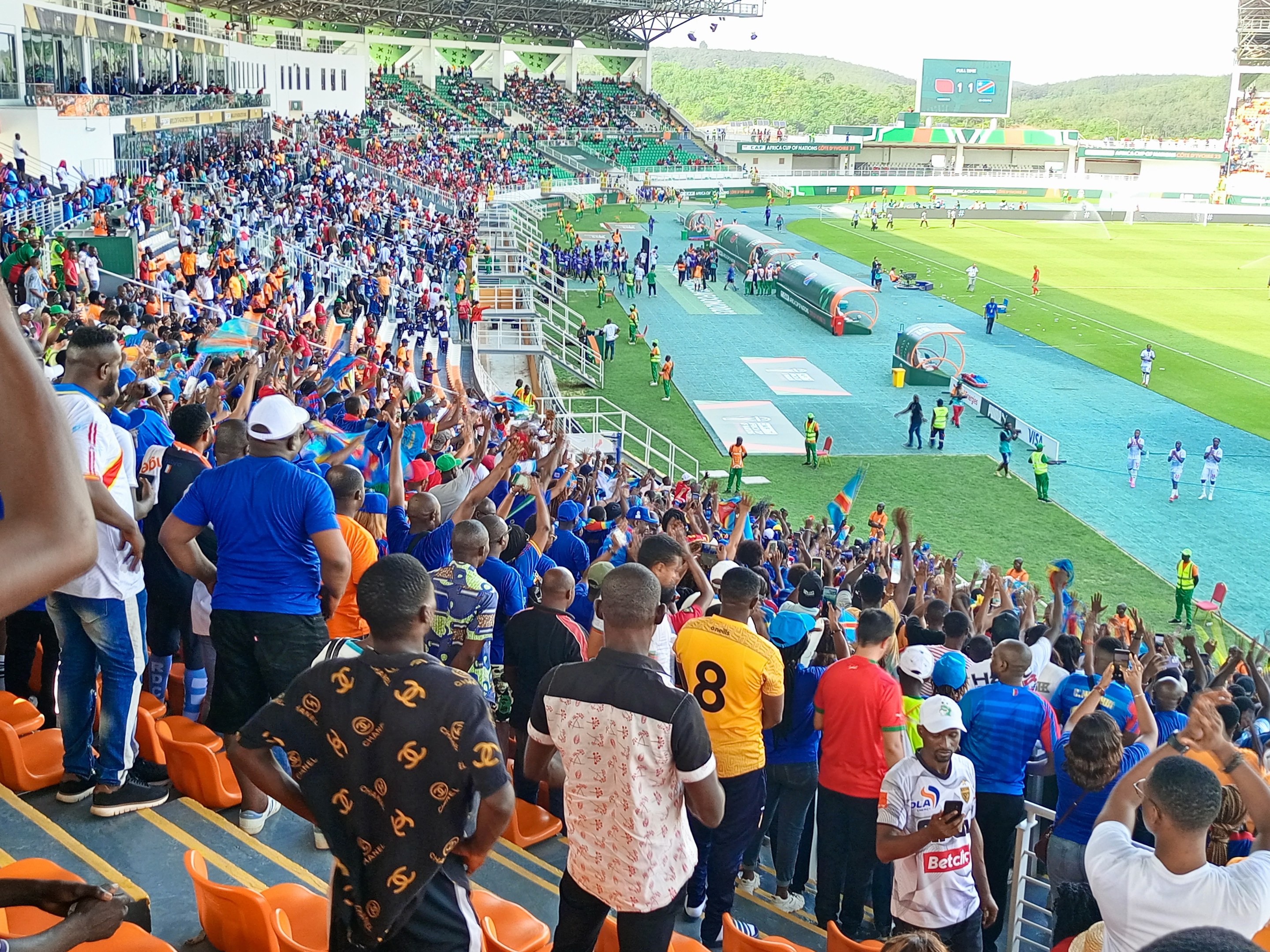 Eine große Menschenmenge steht und sitzt vor einem eingezäunten Fußballfeld mit Hügeln und einem klaren blauen Himmel und wartet auf ein Spiel.