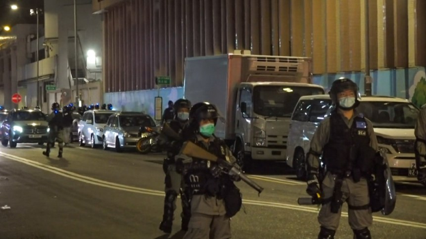 Gruppe von Polizisten in Helmen, Handschuhen und Gesichtsmasken, die auf einer Stadtstraße stehen, einige mit Waffen, mit Fahrzeugen, Laternen, Schildern und Gebäuden im Hintergrund.