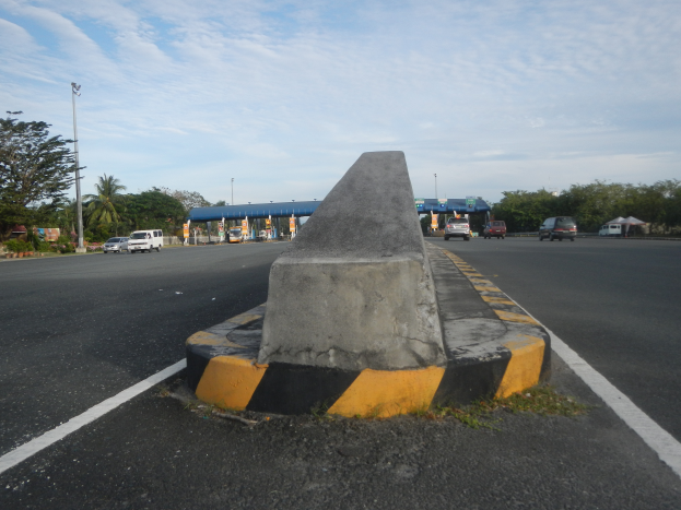 Ein Betonbollard an der Seite einer Straße mit fahrenden Autos, Böumen, Masten, Hütten und einem klaren blauen Himmel im Hintergrund.