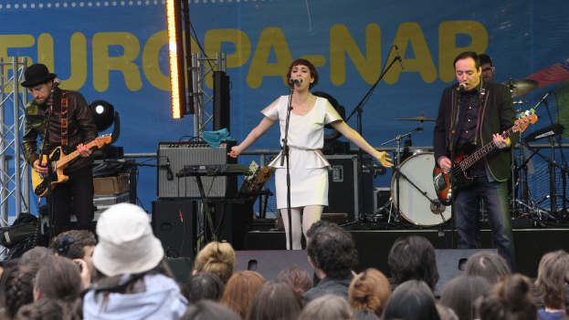 Eine Bühne mit vier Musikern: Eine Frau in einem weißen Kleid singt in der Mitte, zwei Männer spielen Gitarren auf beiden Seiten und eine Menge ist unten mit einer Wand und Lautsprechern im Hintergrund zu sehen.