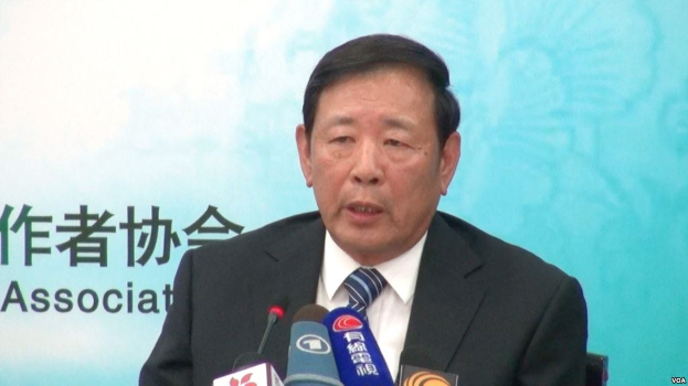 A suited man sits at a table with microphones, speaking to the press, with a banner visible in the background.