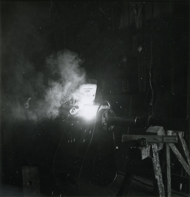 Schwarzes und weißes Foto eines Schweißers, der an einem Metallstück arbeitet, mit Rauch aus der Schweißmaschine und verschiedenen Gegenständen im Hintergrund.