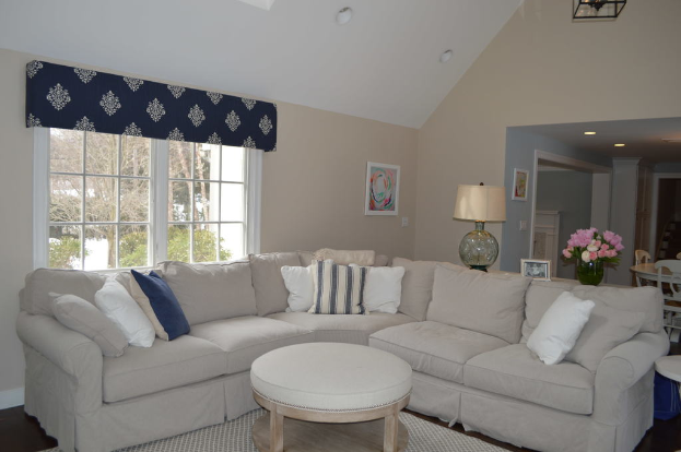 Ein Wohnzimmer mit einer Couch, Kissen, einem Tisch, Stühlen, einem Blumenstrauß, einem Foto, einer Uhr, einem Fenster auf der linken Seite und einer Deckenbeleuchtung.