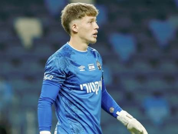 Ein junger Mann in einem blauen Torwarttrikot steht auf einem Fußballfeld, trägt ein blaues T-Shirt und weiße Handschuhe während eines Spiels.