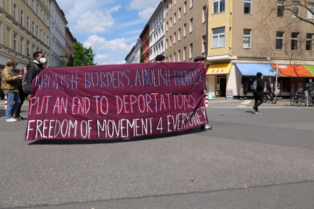 Menschen marschieren mit einem Banner, auf dem "Abolish Borders, Abolish Frontiers, Put an End to Deportations, Freedom of Movement 4 Everyone" steht, durch eine Straße mit Gebäuden, Bäumen und Fahrrädern an der Seite und einer bewölkten Himmel.