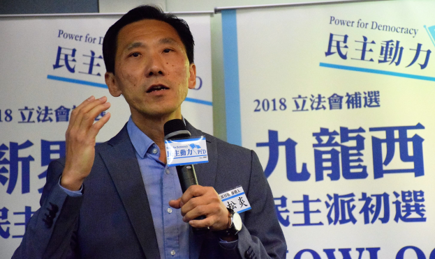 Ein Mann im Anzug, der in ein Mikrofon spricht, vor einem Hintergrund mit Bannern und einer Wand.