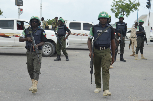 Eine Gruppe von Polizisten in Helmen, die Waffen halten, geht eine Straße entlang mit Fahrzeugen, Laternen, Verkehrsampeln, Texttafeln, Bäumen, Gebäuden und einem bewölkten Himmel im Hintergrund.