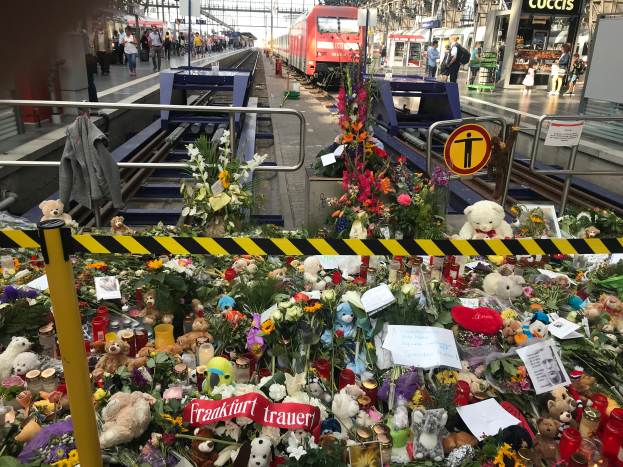 Ein Bahnsteig mit einem Zug im Hintergrund, umgeben von Menschen und verschiedenen Gegenständen wie Teddybären, Blumensträußen und Kerzen, die an ein Unglück in Berlin, Deutschland, erinnern.