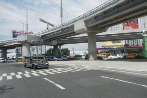 Eine vielbefahrene Hauptstraße mit Fahrzeugen, eine Zebrastreifen, eine Überführung mit Pfeilern, Laternen, Schilder mit Text und Bildern, Bäume, Gebäude und ein bewölkter Himmel im Hintergrund.