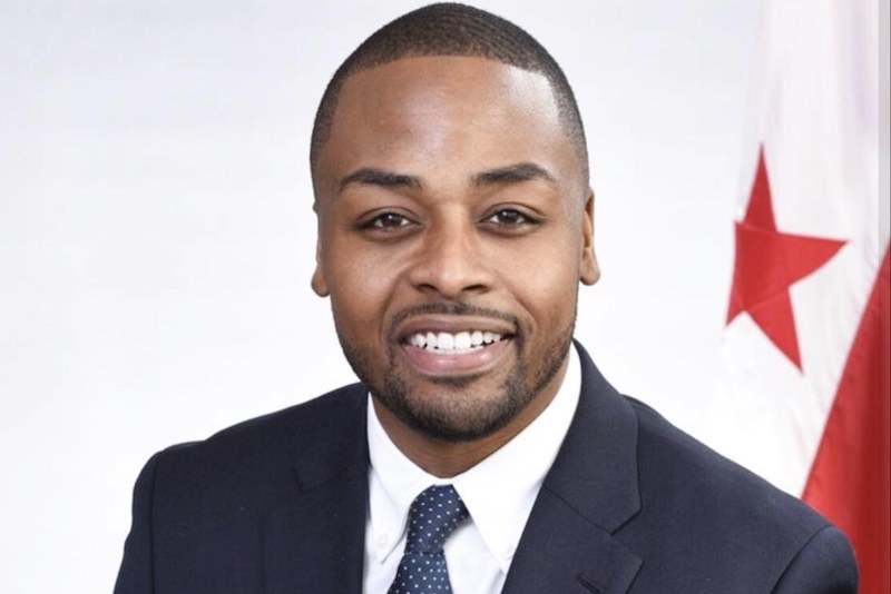 A man in a suit and tie smiles at the camera with a red and white flag and text indicating he is the newly appointed UN ambassador in the background.
