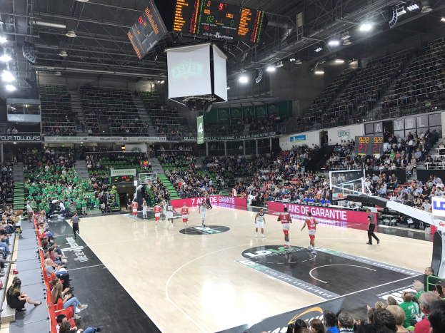 Basketball game in a large arena with a crowd, scoreboards displaying "Asvel vs Galatasaray," and overhead lighting and scoreboard.
