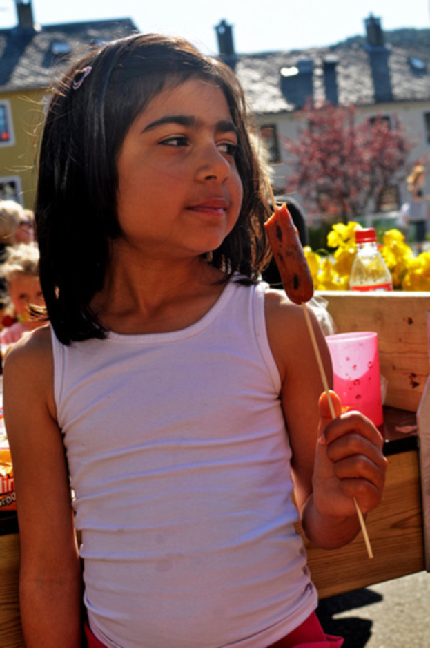 Ein Kind, das ein Lebensmittel hält, mit Häusern, Bäumen, anderen Kindern, Blumen, einer Flasche und Tabletts mit verschiedenen Gegenständen im Hintergrund.