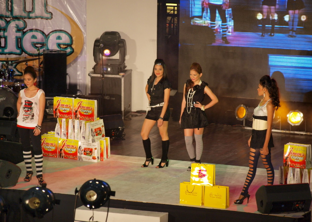 Vier Frauen in einem Raum mit verschiedenen Gegenständen, Taschen und Equipment wie Trommeln, Lautsprechern, Lichtern, einem Bildschirm und einem Turm, mit Text auf einer Tafel im Hintergrund.