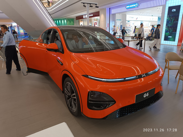 Oranger Elektroauto in einer Ausstellungshalle umgeben von Menschen, mit Stühlen und einem Wagen rechts und Geschäften im Hintergrund.