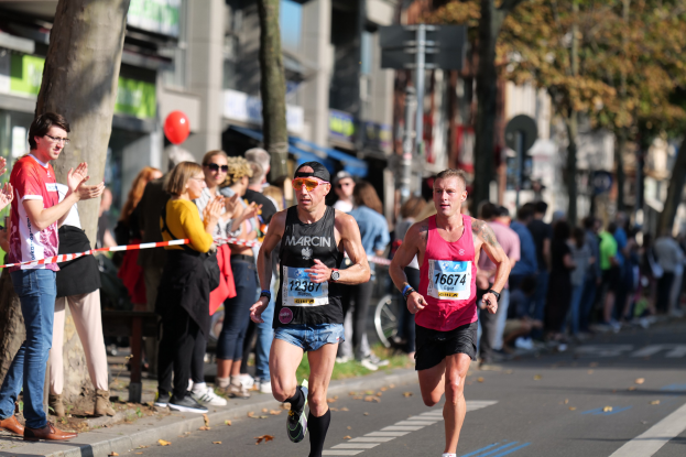 Eine Gruppe von Marathonläufern auf einer baumbestandenen Straße, einige tragen Kappen und Schutzbrillen, andere klatschen; im Vordergrund ist eine Person mit Rucksack und Turnschuhen zu sehen.