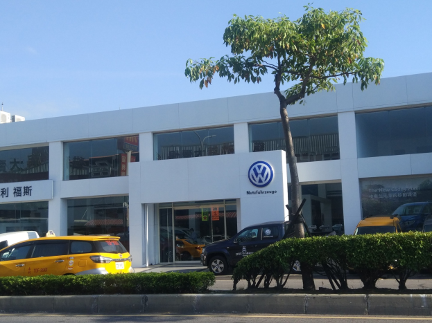Außenansicht einer Volkswagen-Niederlassung mit parkenden Autos davor, umgeben von Pflanzen und einem Baum, mit Glaswänden und -fenstern unter einem sichtbaren Himmel.