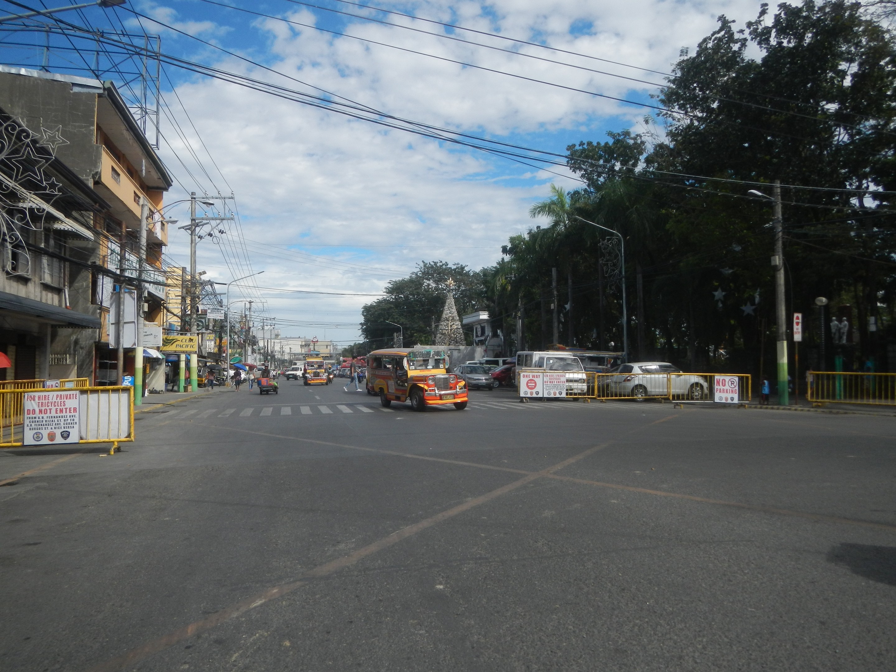 A busy city street with cars, buses, trucks, barricades, signboards, light poles, electric poles with wires, buildings, trees, and a cloudy sky.