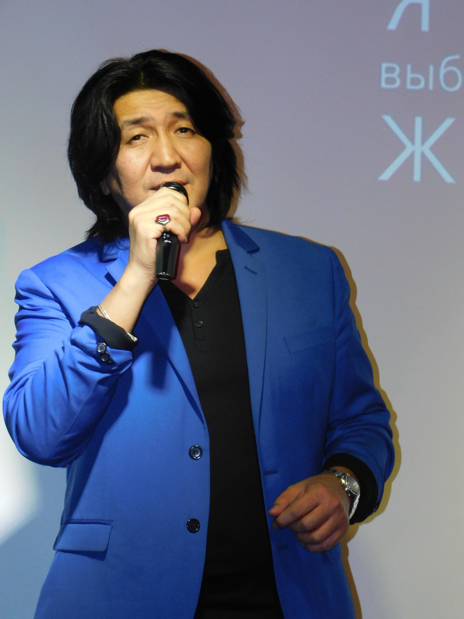 Ein Mann in einem blauen Anzug und schwarzem Hemd hält ein Mikrofon und steht vor einem Bildschirm mit Text, mit einem ernsten Gesichtsausdruck.