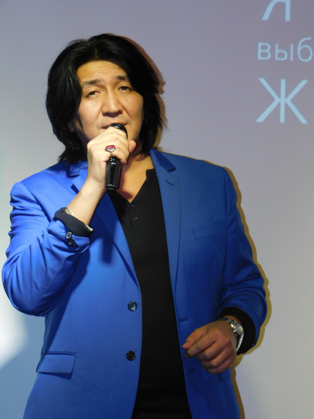 Ein Mann in einem blauen Anzug und schwarzem Hemd hält ein Mikrofon und steht vor einem Bildschirm mit Text, mit einem ernsten Gesichtsausdruck.