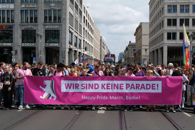 Eine Gruppe von Menschen marschiert auf einer Straße in Berlin mit einem pinken "Happy Pride March"-Schild, umgeben von Gebäuden, Laternenmasten und Verkehrszeichen unter einem bewölkten Himmel.