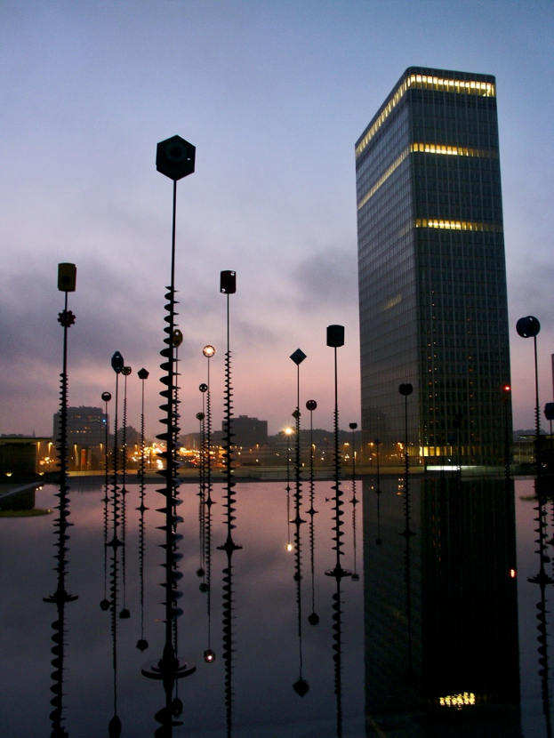Mehrere Masten und Gebäude mit Lichtern, unter einem bewölkten Himmel, mit ihren Spiegelungen.