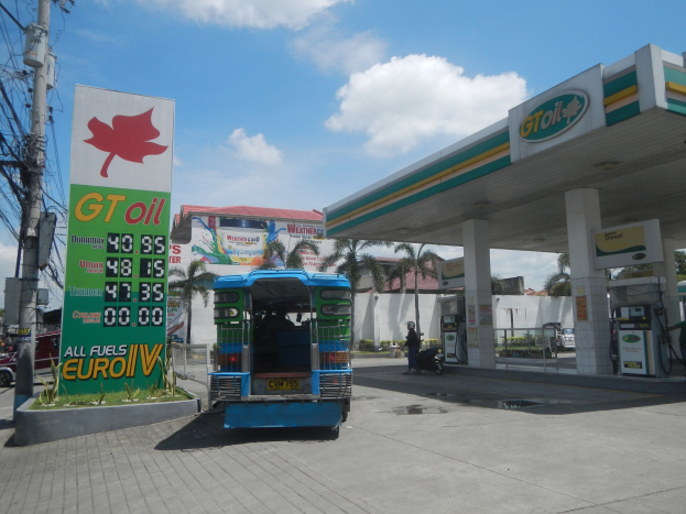 Tankstelle mit einem LKW davor, Fahrzeugen auf der Straße, einem Radfahrer, Zapfsäulen, Bäumen, Gebäuden, Wolken und einem Strommast mit Drähten.