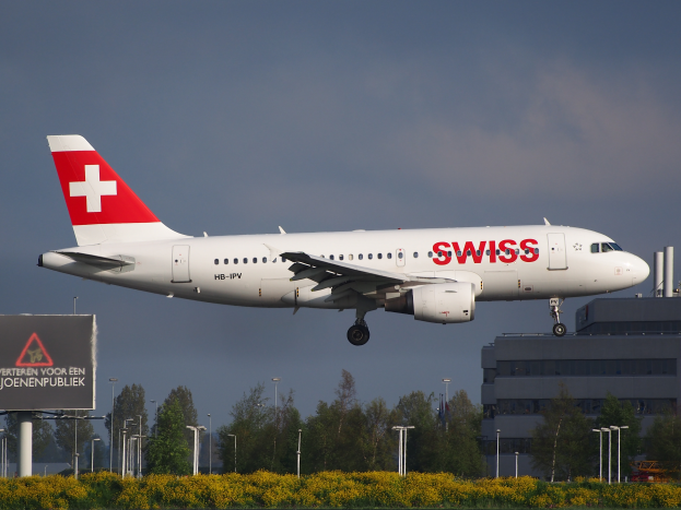 Ein Airbus A320-200 der Swiss Airlines startet am Frankfurter Flughafen, umgeben von Grünflächen und einem Gebäude sowie Lichtmasten im Hintergrund, unter einem bewölkten Himmel, mit einer Texttafel im Vordergrund.