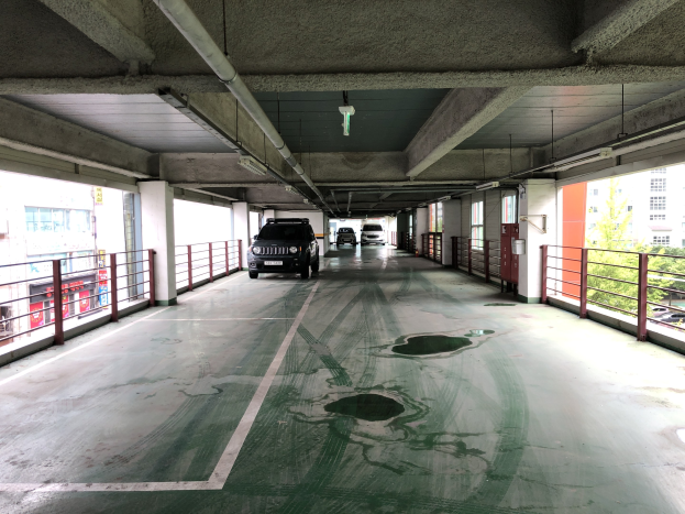 Ein mehrgeschossiges Parkhaus mit parkenden Autos, Geländern, Säulen, Deckenleuchten, freiliegenden Rohren und umgebenden Bäumen, vor einem Hintergrund von Gebäuden mit Fenstern in einer Stadtlandschaft.