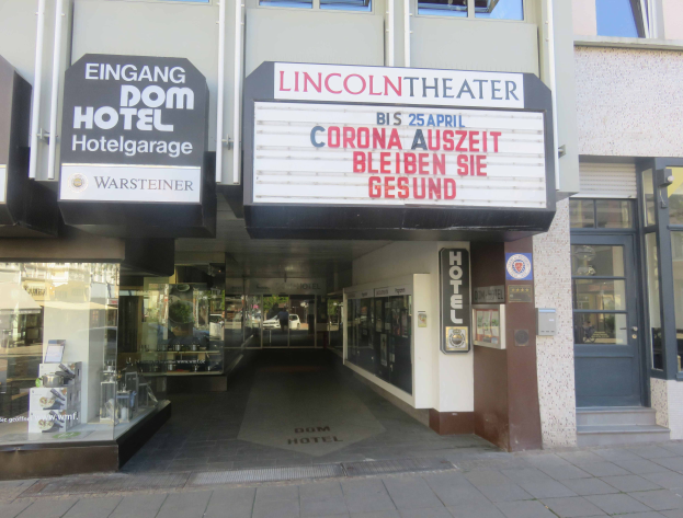 Das Lincoln Theater in Berlin, Deutschland, ein Gebäude mit Glasfenstern und -türen sowie einer Tafel mit Text darauf, das verschiedene Gegenstände im Inneren zeigt und den Eindruck einer pulsierenden Stadtlandschaft vermittelt.