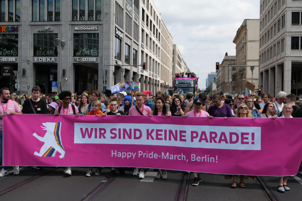 Eine Gruppe von Menschen marschiert auf einer Berliner Straße mit einem pinken Banner, auf dem "Happy Pride March" steht, vorbei an Gebäuden, Laternenpfählen und Verkehrsampeln.