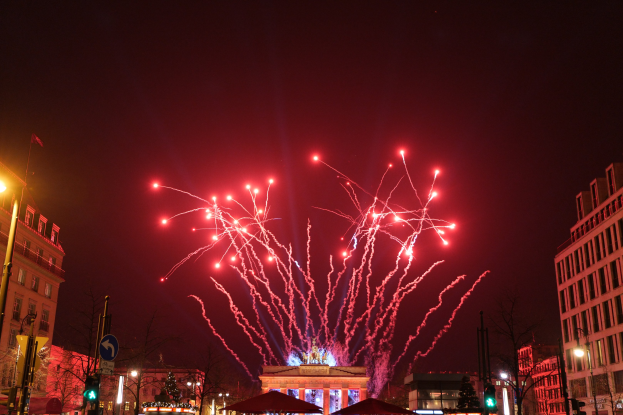 Eine belebte Stadtstraße bei Nacht am Silvesterabend in Berlin, mit Gebäuden, Bäumen, Laternenmasten, Ampeln, Schildern, Zelten, Menschen und einem prächtigen Feuerwerk, das den Himmel erhellt.