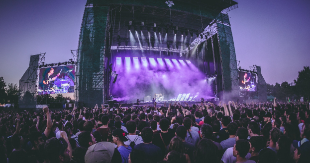 Eine große Menschenmenge auf einem Musikfestival schaut auf einer beleuchteten Bühne mit Instrumenten und Bildschirmen, die vor einem Gebäude, Bäumen und einem blauen Himmel stehen.