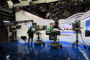 Ein Mann steht vor einem Fernseher und hält ein Mikrofon, mit Kameras auf dem Boden, einem Stuhl, einem Bildschirm, einer Wand und einer Decke mit Deckenleuchten, was auf eine Live-Übertragung hindeutet.