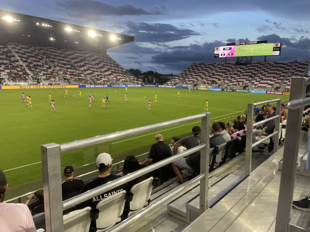 A soccer game between Orlando City SC and Philadelphia Union in a stadium with spectators in the stands, scoreboard visible, surrounded by trees under a cloudy sky.