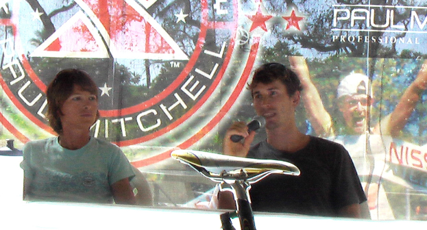 Eine Gruppe von Menschen auf Stühlen sitzend, ein Mann mit einem Mikrofon in der Hand, ein Banner mit einer Person mit einer Mütze und ein Fahrradsattel im Vordergrund.